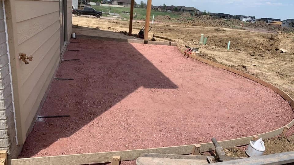 A graded, compacted base pad beside a garage with a wood form set along the edge