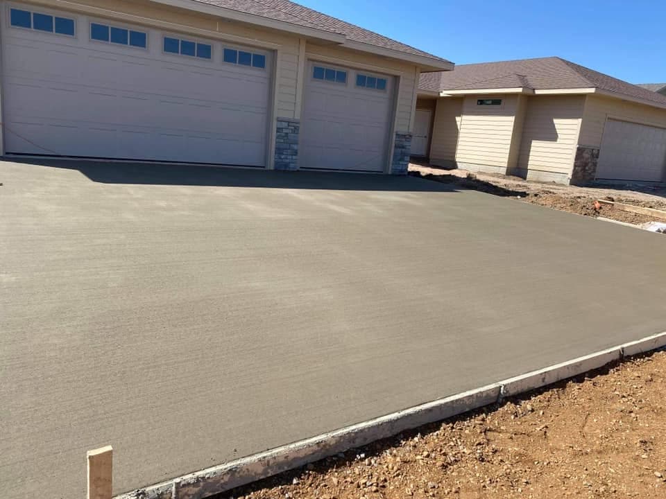 A broad broom-finished concrete driveway freshly poured in front of a two-stall garage on a new home, with a wood-formed edge and graded ground alongside