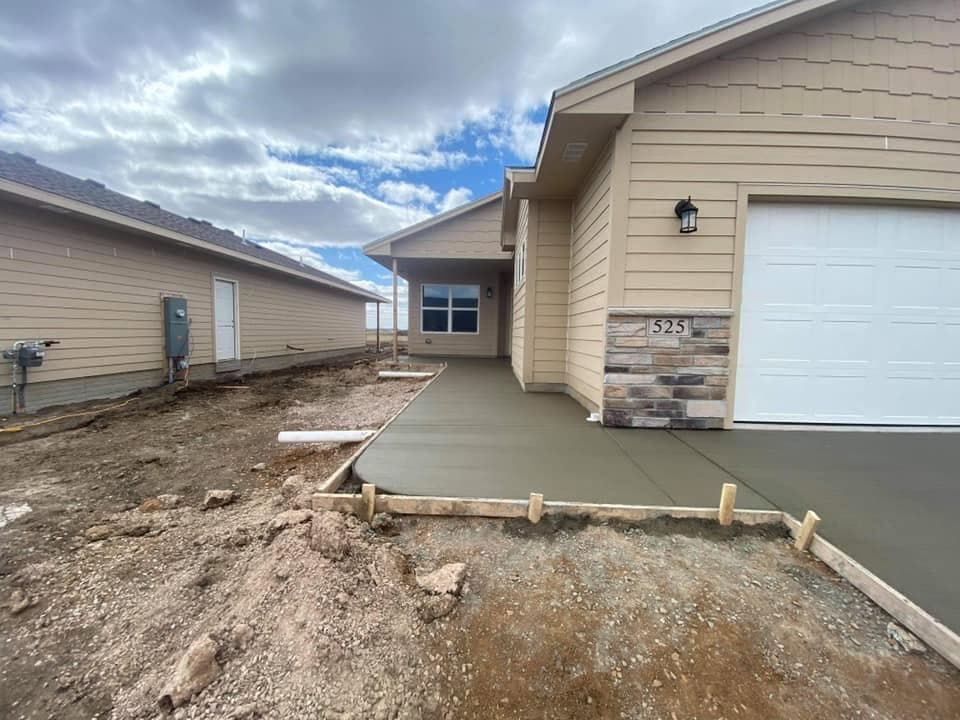 A freshly finished concrete approach and front walk beside the garage of a new home, wood forms set along the graded dirt