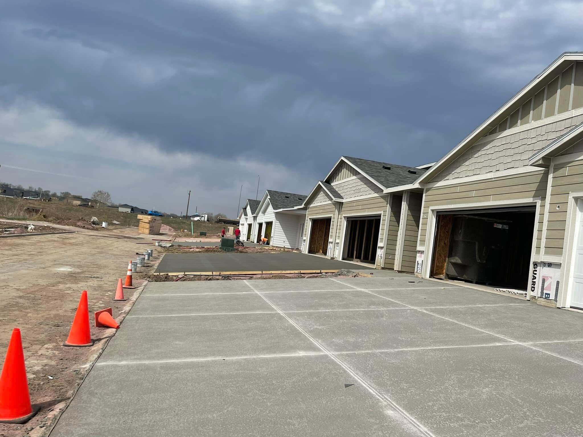 A cured driveway apron with saw-cut control joints running along a row of new townhome garages, the next pour going down behind it