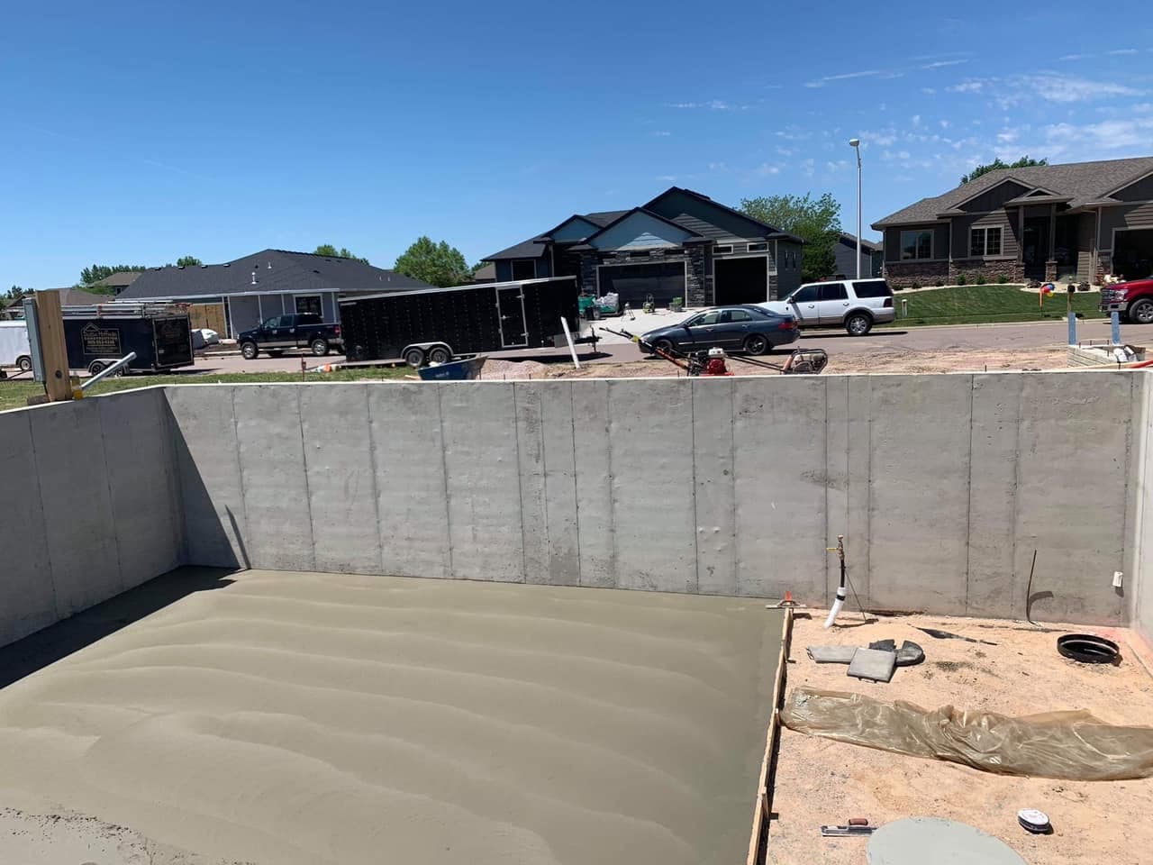 A freshly floated basement floor slab poured inside tall concrete foundation walls, with a plumbing rough-in set and a trowel resting at the edge