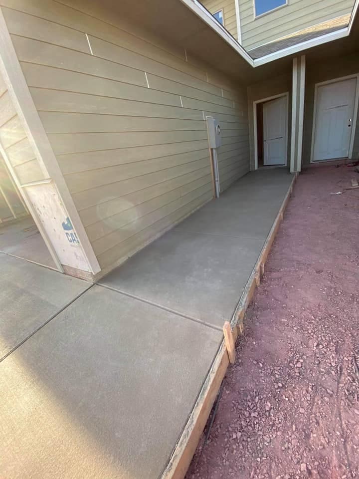 A finished concrete walk and apron running along the side of a new home to the entry doors, with a formed edge along the graded ground