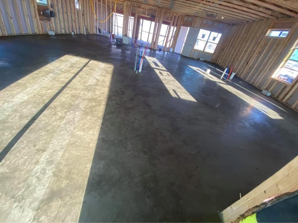 A large hard-troweled concrete shop floor inside a framed building, plumbing stub-ups set and sunlight across the fresh finish