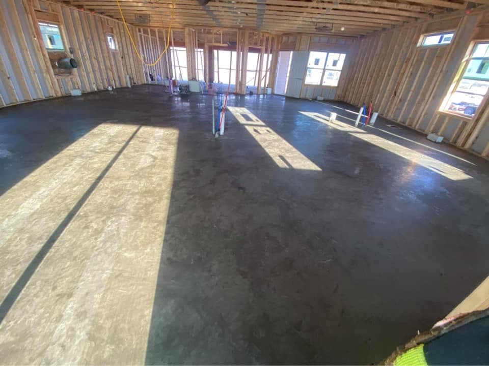 A large hard-troweled concrete floor inside a newly framed building, plumbing stub-ups set and sunlight falling across the fresh finish