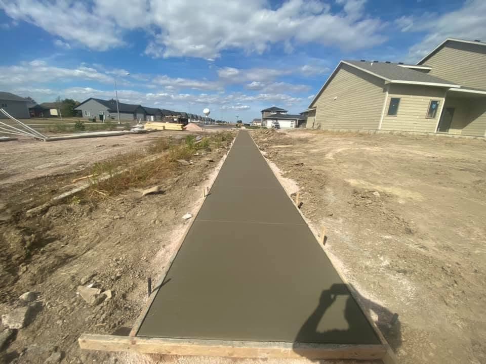 A long straight freshly poured concrete sidewalk running between graded lots toward the horizon, with wood-formed edges