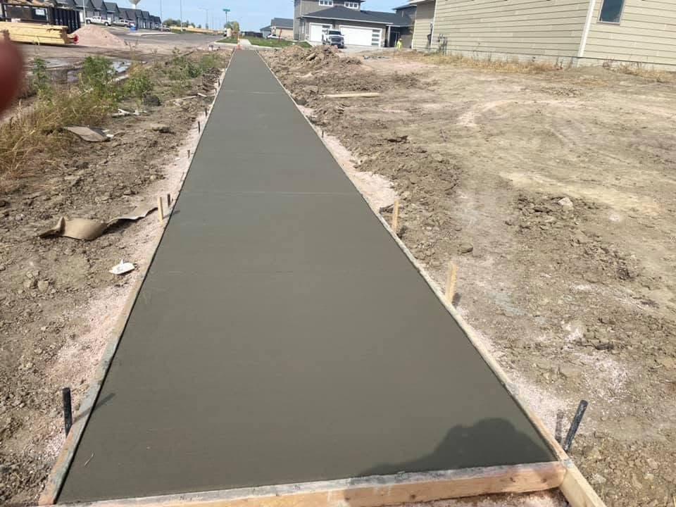 A long straight freshly poured concrete sidewalk with formed edges running toward a row of garages